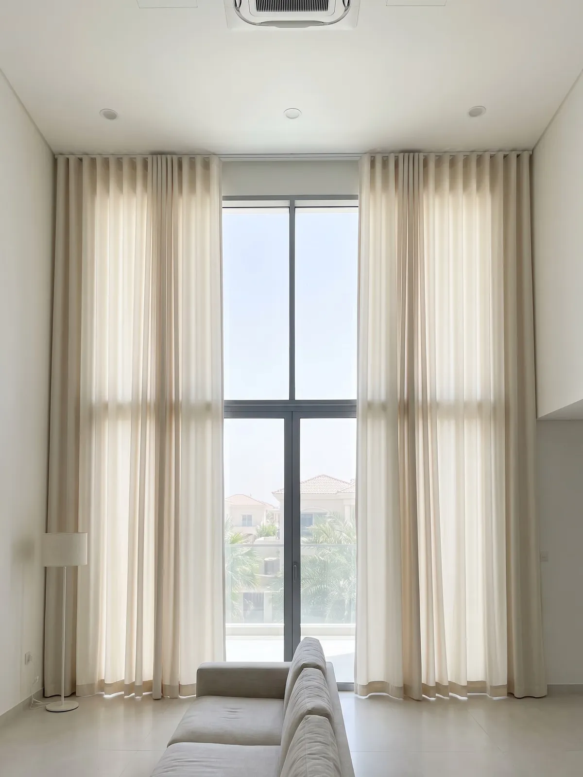 Cream wave curtains drawn open either side of a two-storey window onto villa roofs and palms, a floor lamp at left, a sofa below