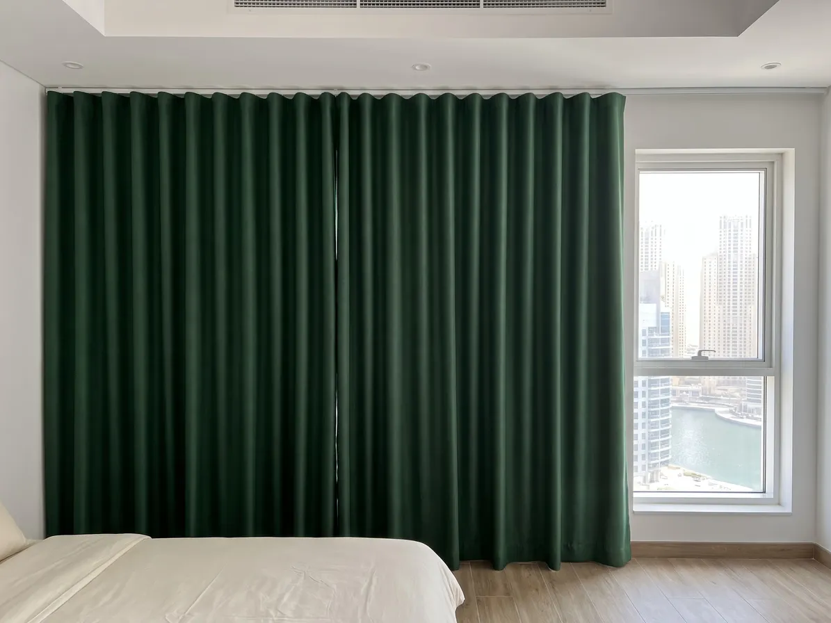 Deep green wave curtains closed on a ceiling track across a bedroom wall, a window with a marina view at right, a bed at lower left