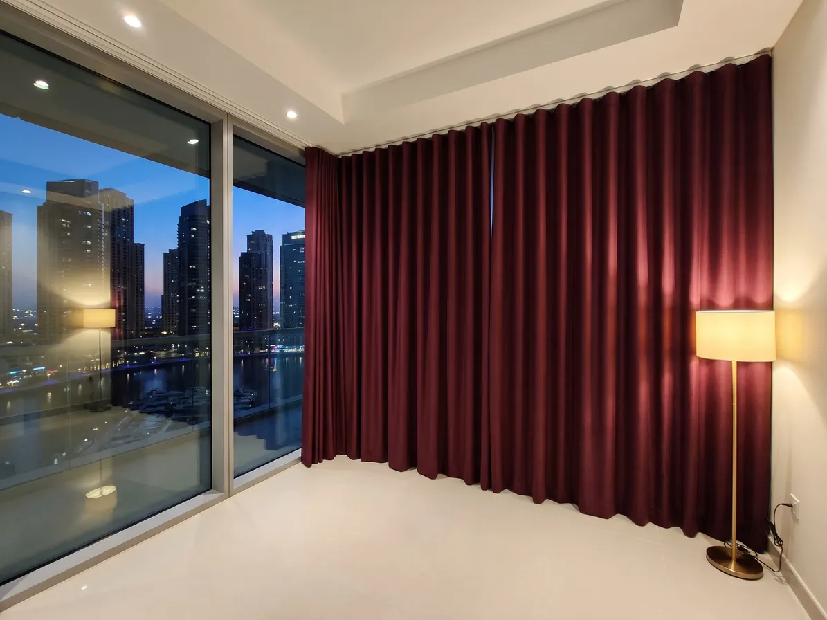 Burgundy wave curtains closed along the corner of a glass wall at dusk, marina lights through the glass at left, a lit floor lamp at right