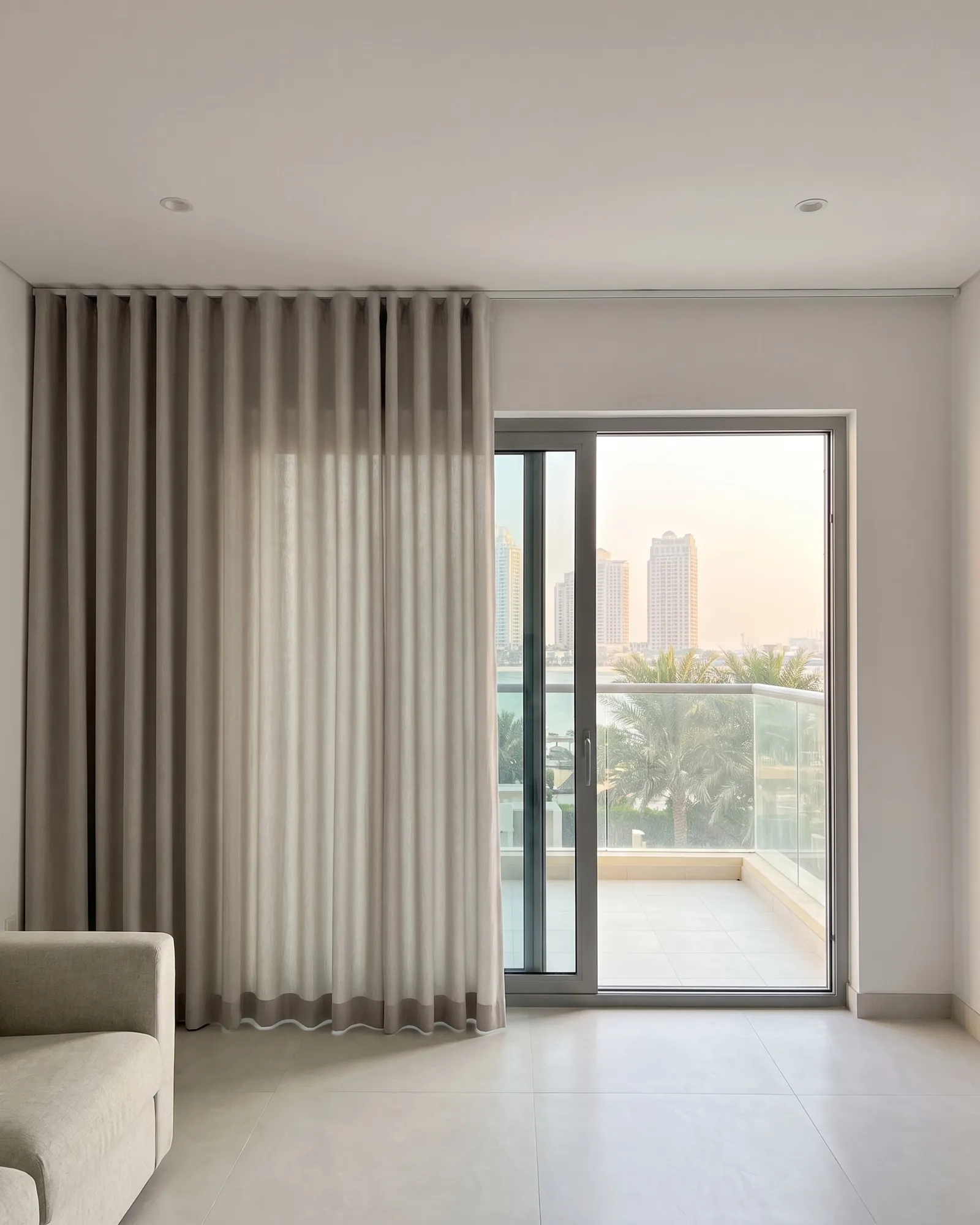 Greige wave curtain half drawn beside a sliding balcony door, towers, palms and sea beyond, a sofa arm at lower left