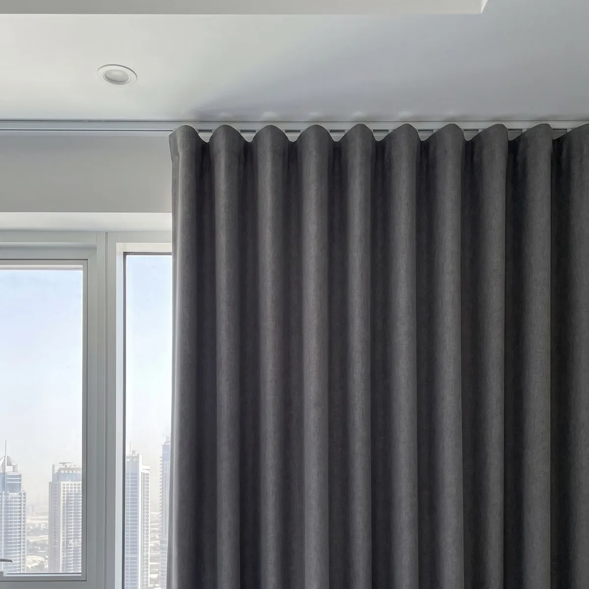 Charcoal-grey wave heading hanging from a white ceiling track, evenly spaced slots along the track above each wave, a window at left