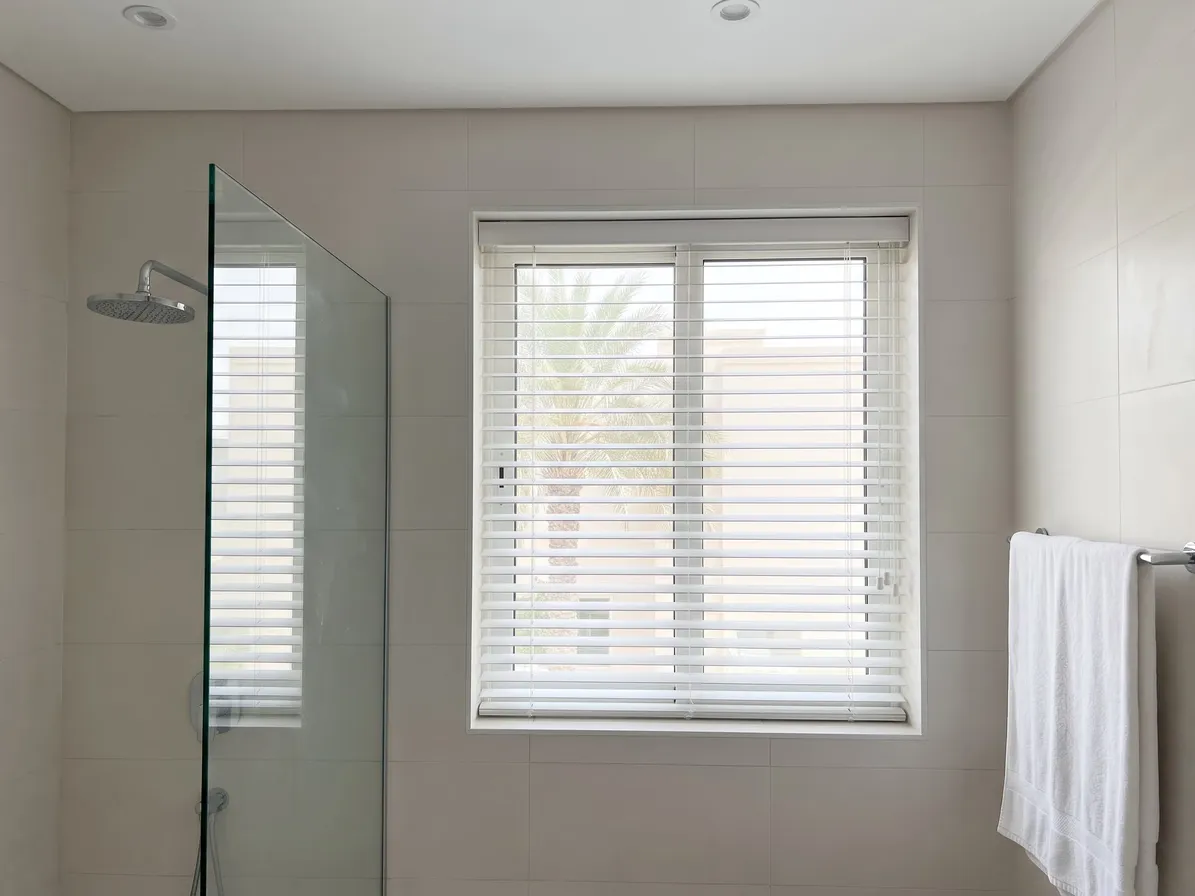White faux-wood blind with tilted slats in a bathroom window, a glass shower screen at the left and a white towel on a rail at the right