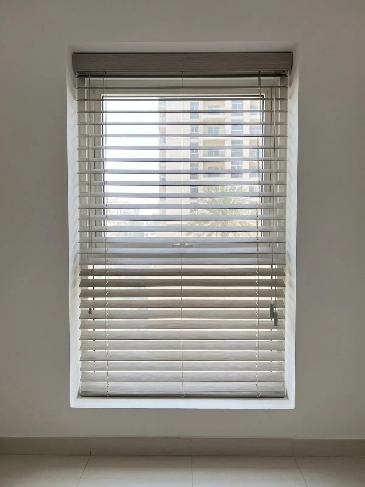 Limed grey oak faux-wood blind with tilted slats on thin ladder strings, a tower and palms between the slats, cords with tassels at the right
