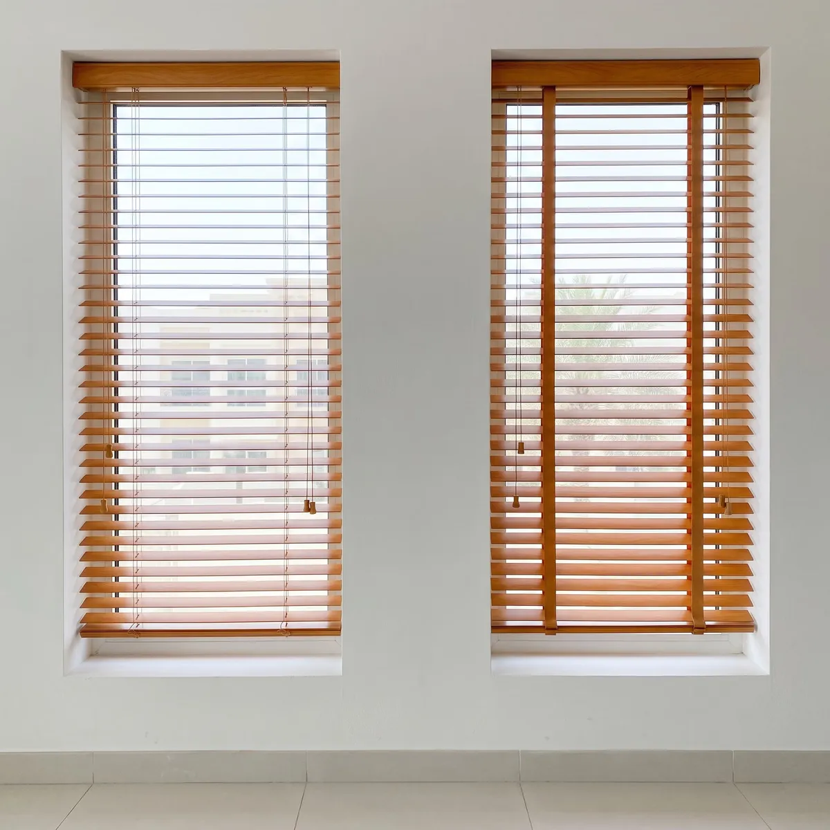 Two maple faux-wood blinds on tall windows side by side, the left one on thin strings and the right one on wide cloth tapes