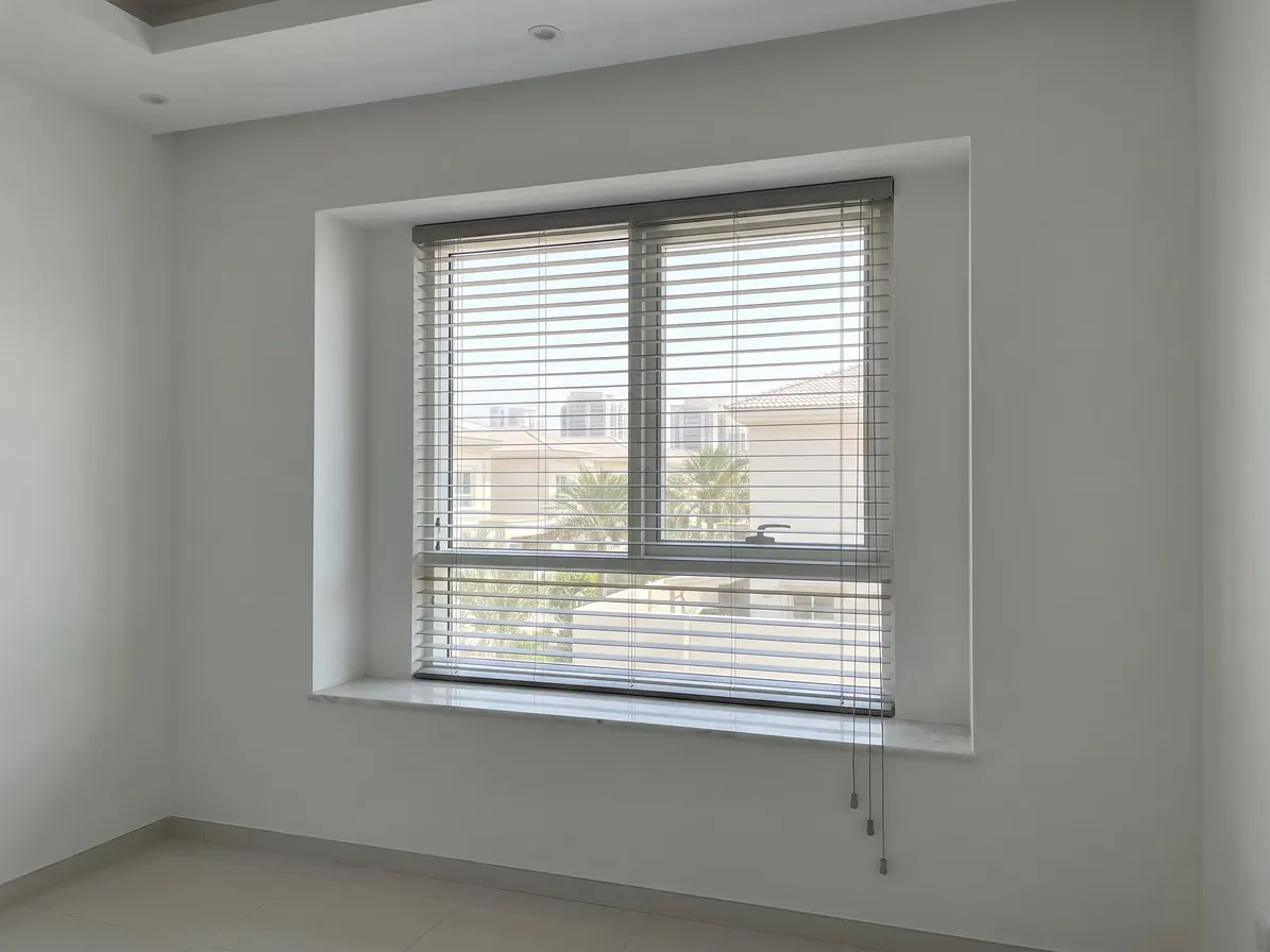 Pebble grey faux-wood blind inside a deep window recess with a wide sill, villas and palms between the slats, cords hanging at the right