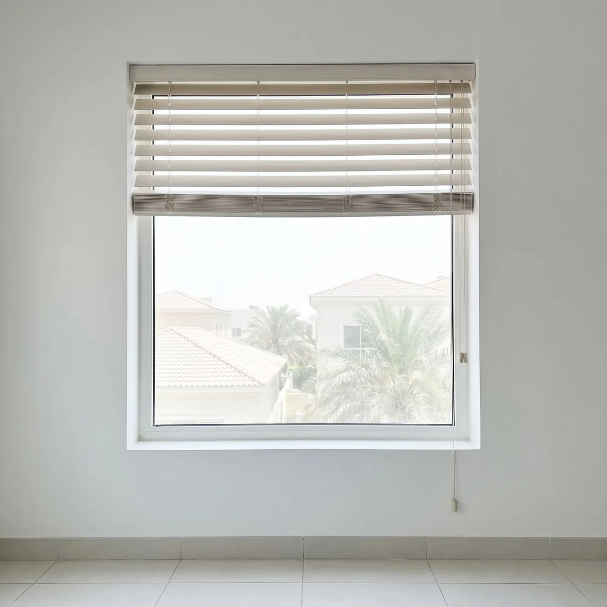 Whitewashed beige faux-wood blind pulled up under its valance, villa roofs and palms in the clear glass below, its pull cord hanging at the right