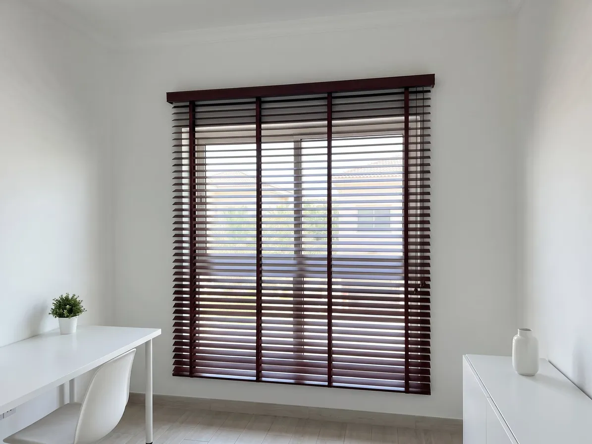 Mahogany faux-wood blind fixed outside a study window, past the frame, a white desk with a plant at the left and a cabinet at the right
