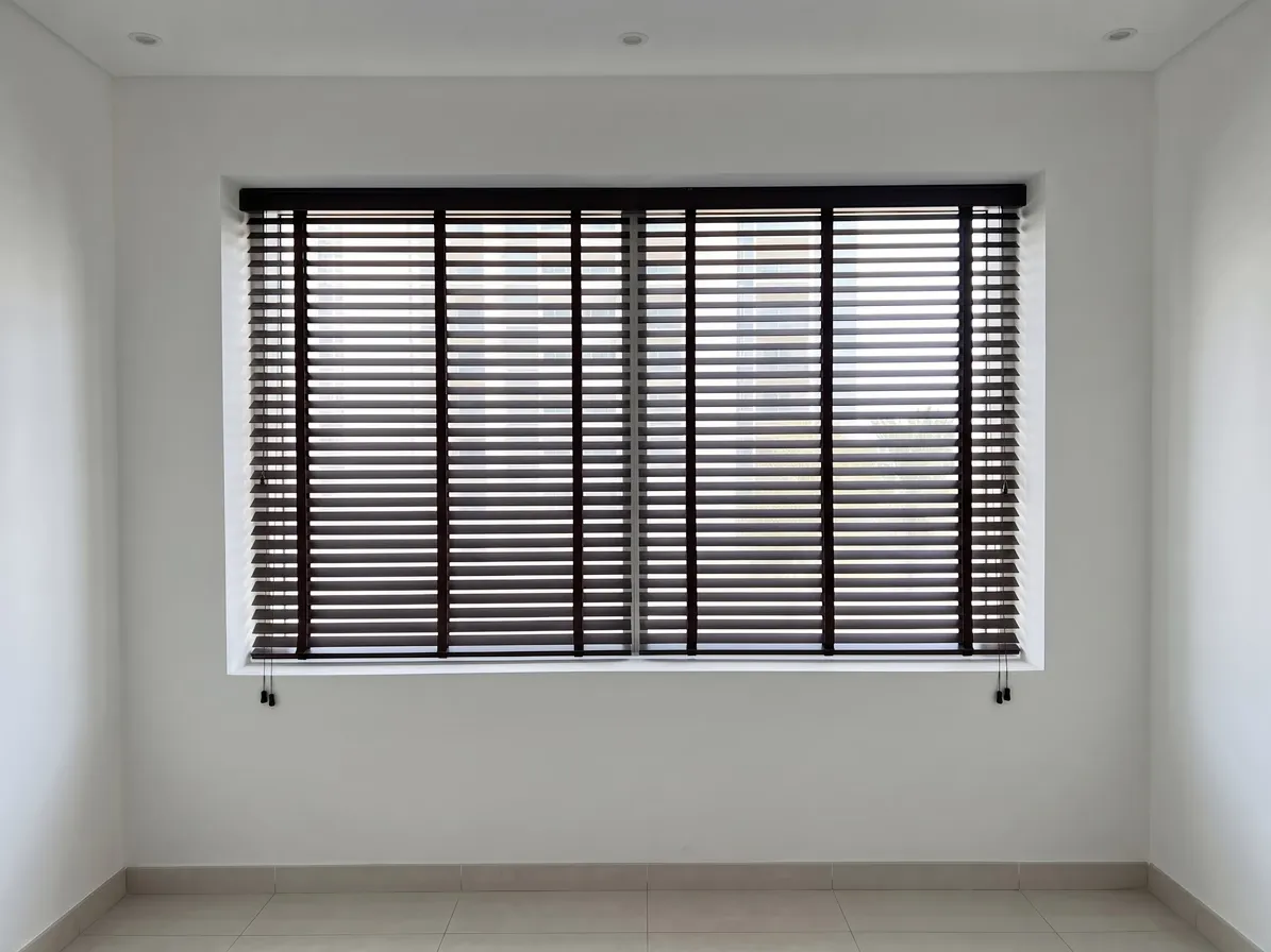 Two dark wenge faux-wood blinds side by side across one wide window, slats tilted part-open, cords with small tassels at the outer ends