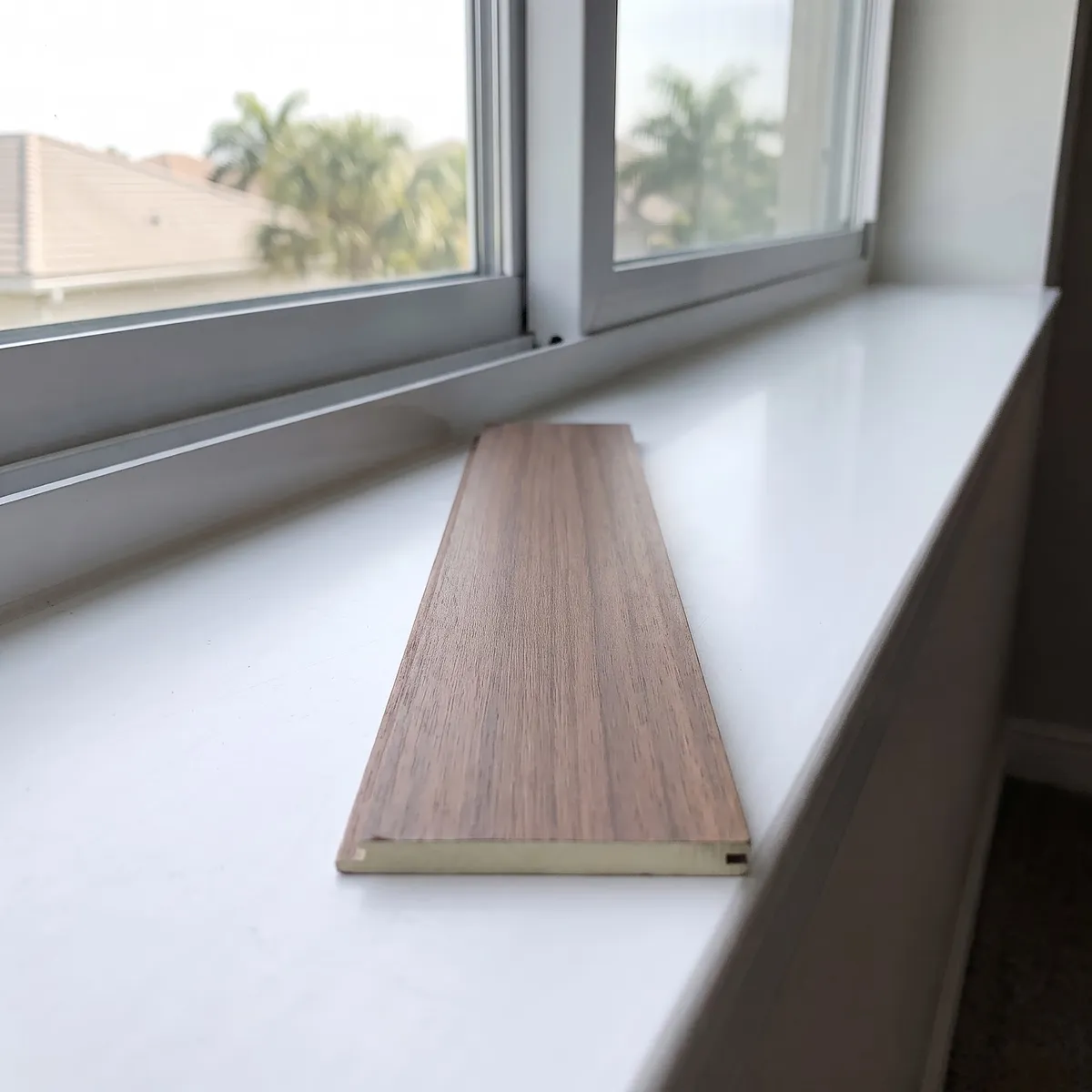 A walnut faux-wood slat lying on a window sill, its cut end showing a pale even core, palms and a villa roof through the glass