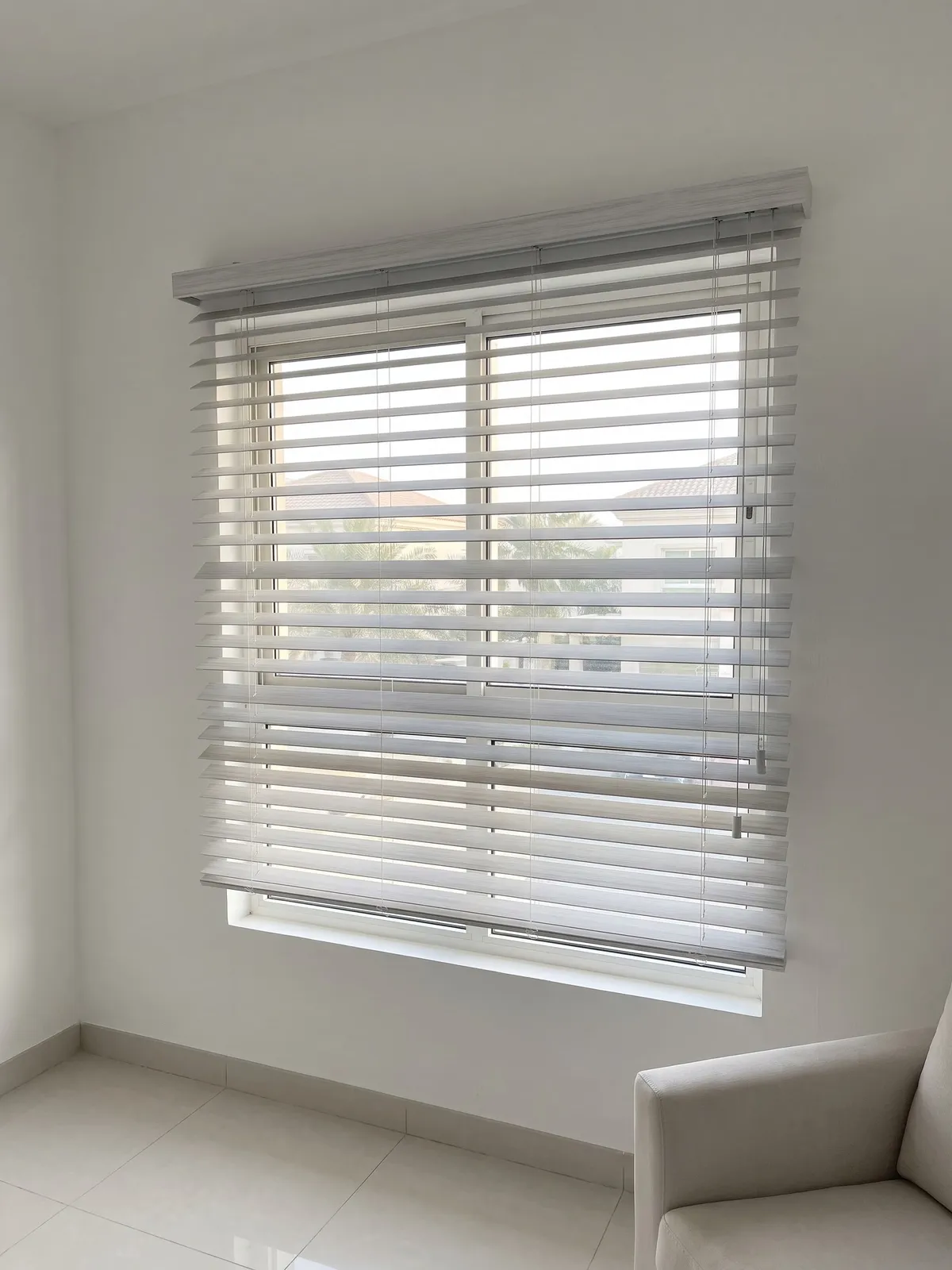 Whitewashed grey faux-wood blind with tilted slats over a window, cords with tassels at the right and a pale armchair at the lower right