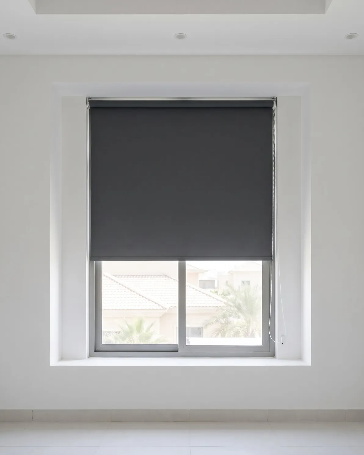 Charcoal roller blind lowered over the top two thirds of a window inside a deep recess, villa roofs and palms below it