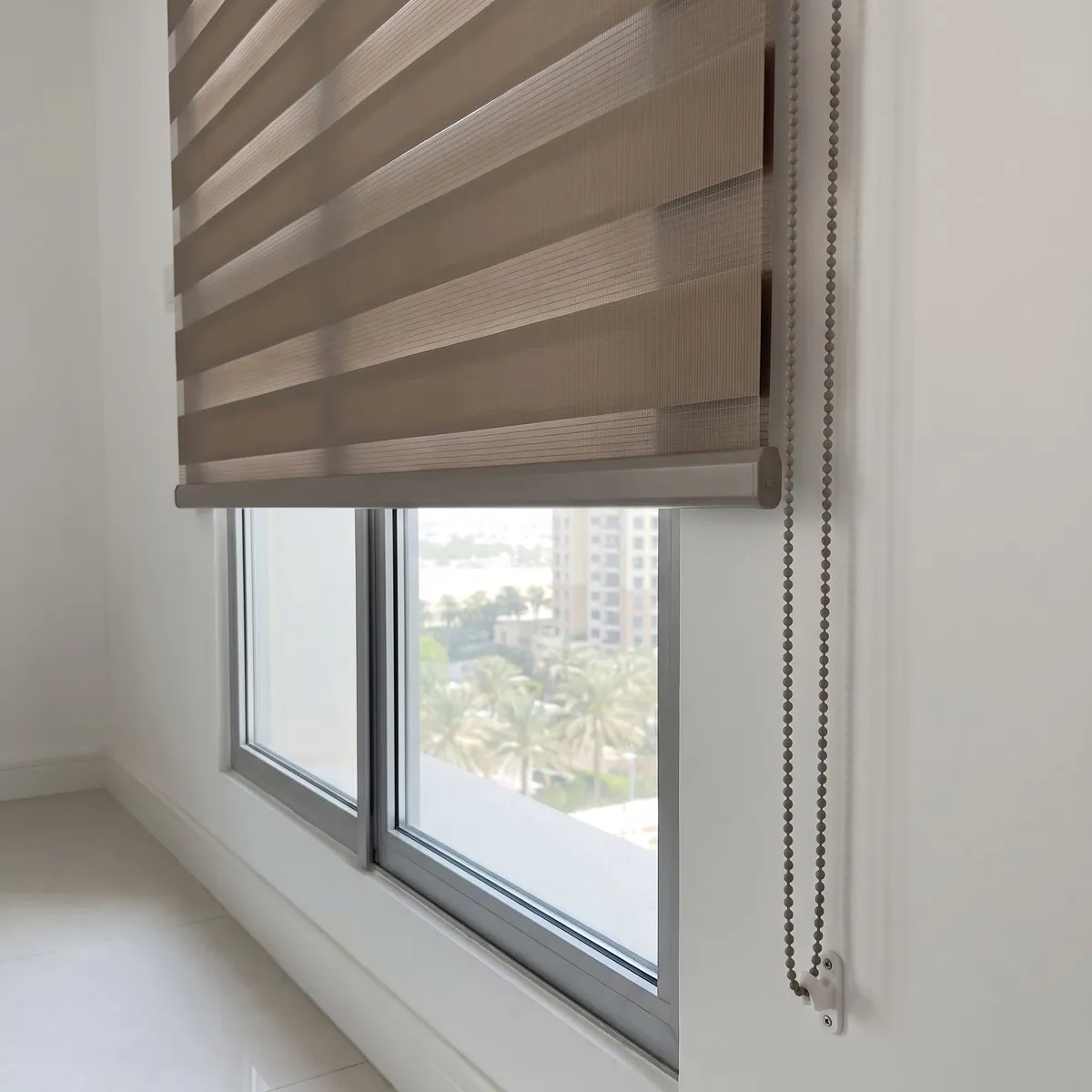 Taupe zebra blind seen from the side, both fabric layers meeting its bottom rail, a bead chain at the right, palms through the glass below