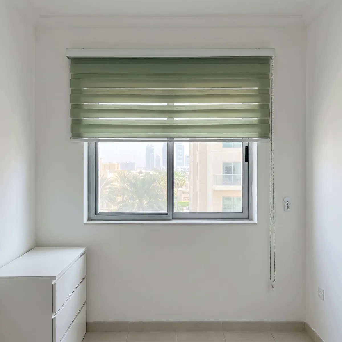 Muted sage zebra blind raised halfway on a window, its chain held by a small clip on the wall at the right, a white chest below