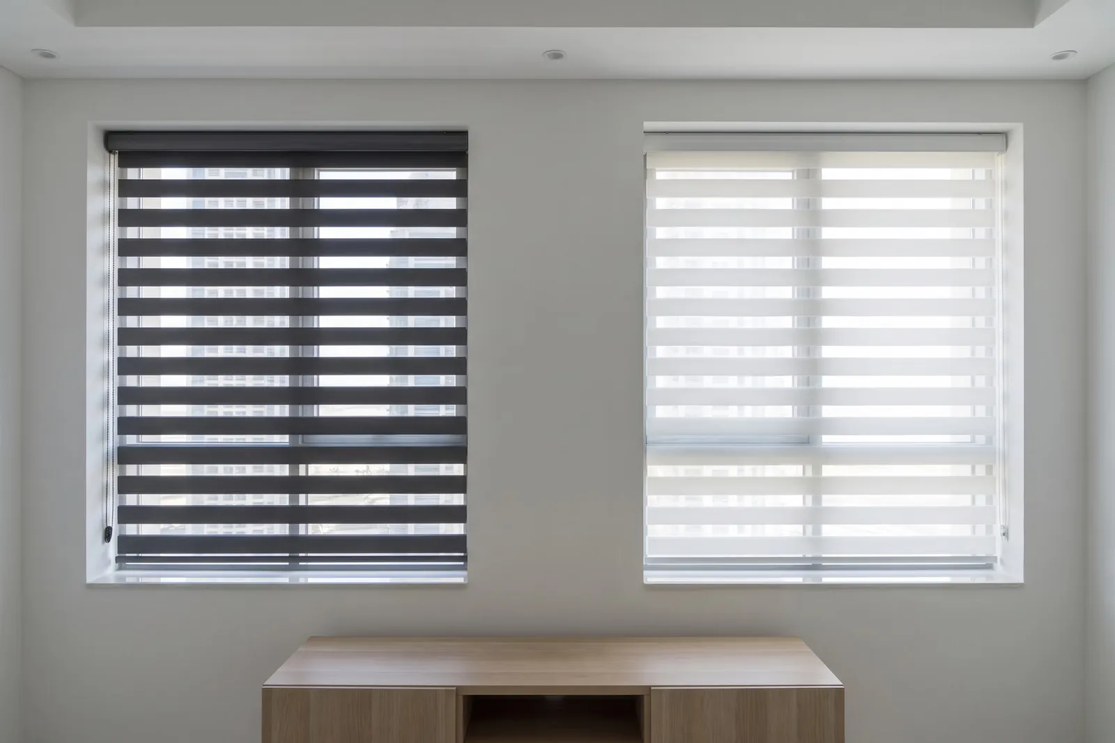 Charcoal and white zebra blinds on two matching windows side by side, in the same daylight, a pale wood sideboard below them