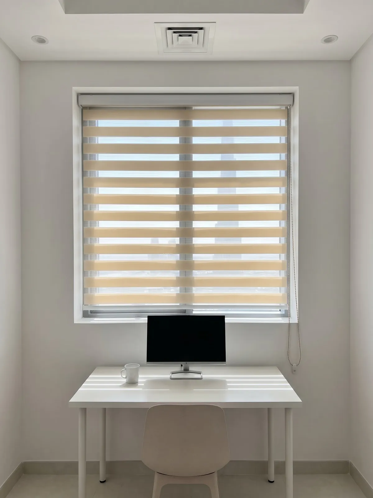Sand zebra blind on a study window with its clear stripes narrowed, a white desk with a monitor and a chair below it