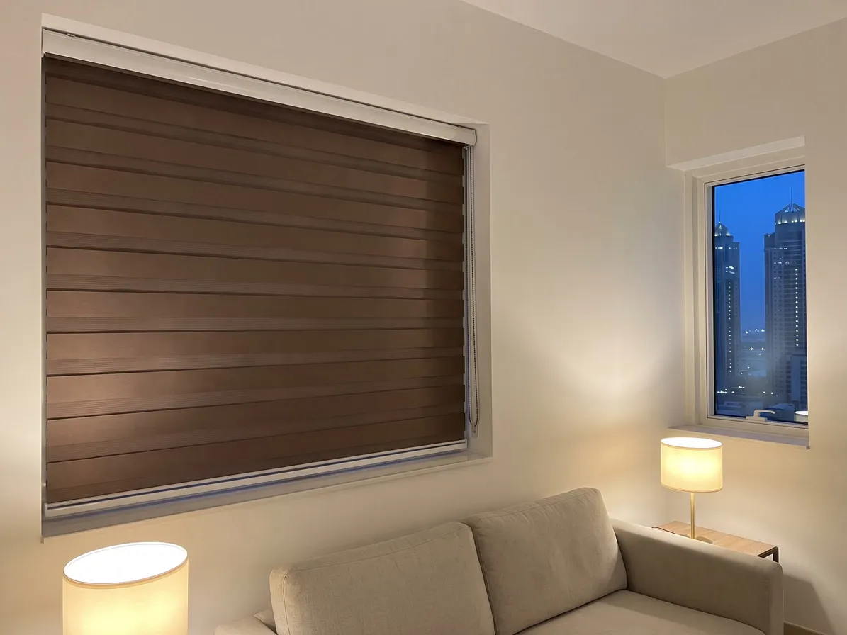 Mocha brown zebra blind closed on a living-room window at dusk, two lamps lit, a beige sofa, lit towers in a narrow window