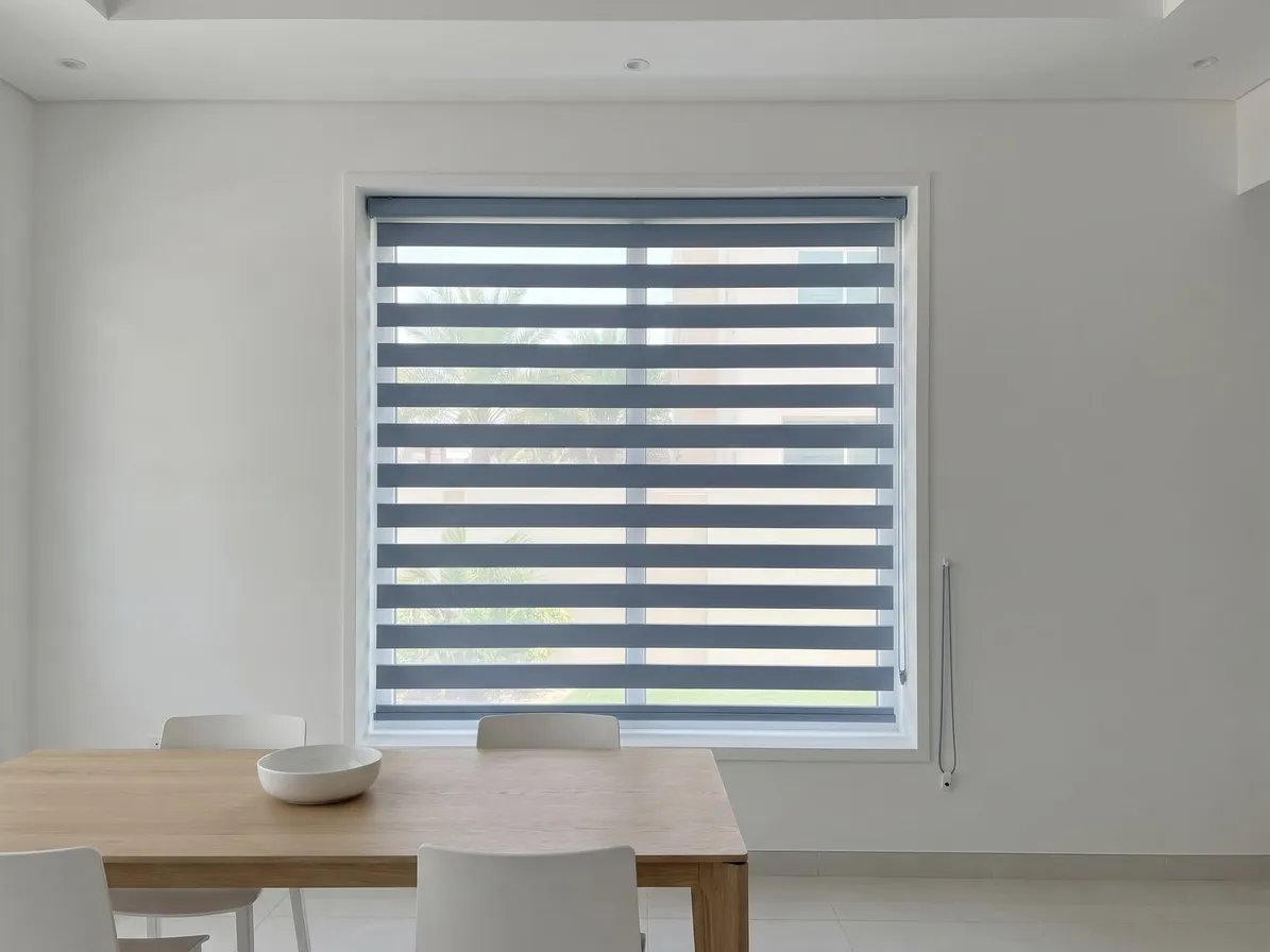 Dusty blue zebra blind inside a dining-room window recess with its stripes open, a wooden table with a white bowl and chairs