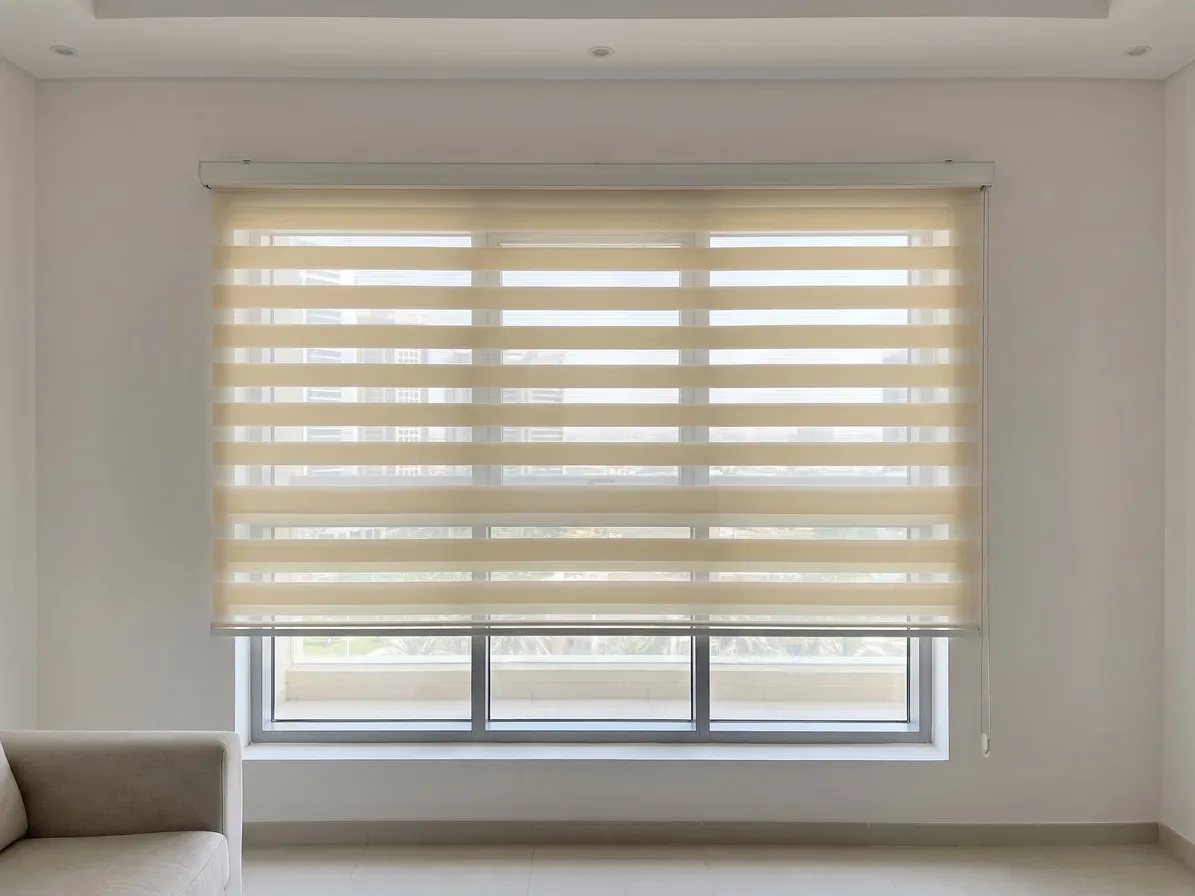 Cream zebra blind fixed on the wall above a living-room window and running past its frame, stripes open, a sofa at the left