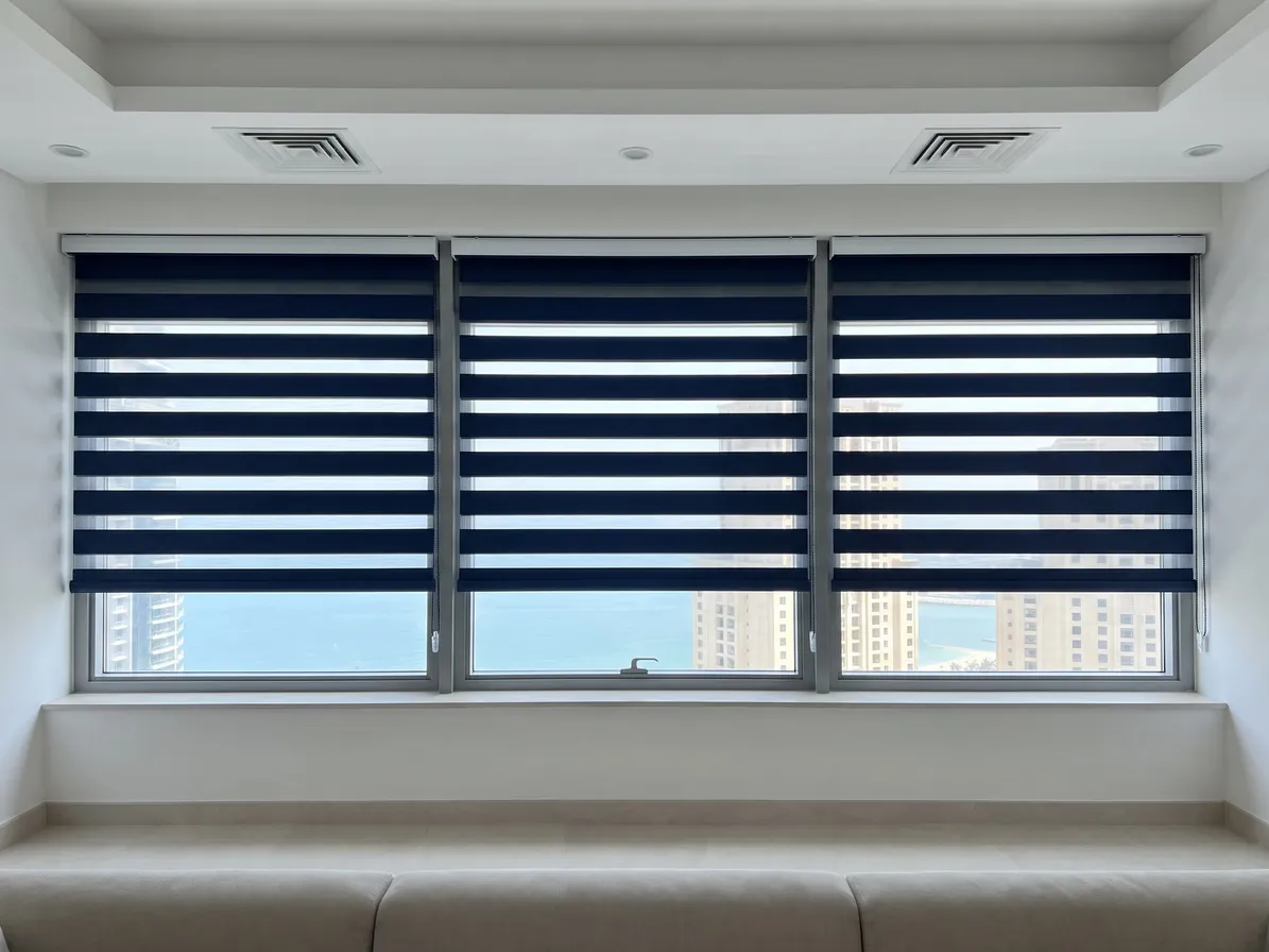 Three navy zebra blinds across one wide window with their stripes lined up, the sea and towers below, a window seat in front