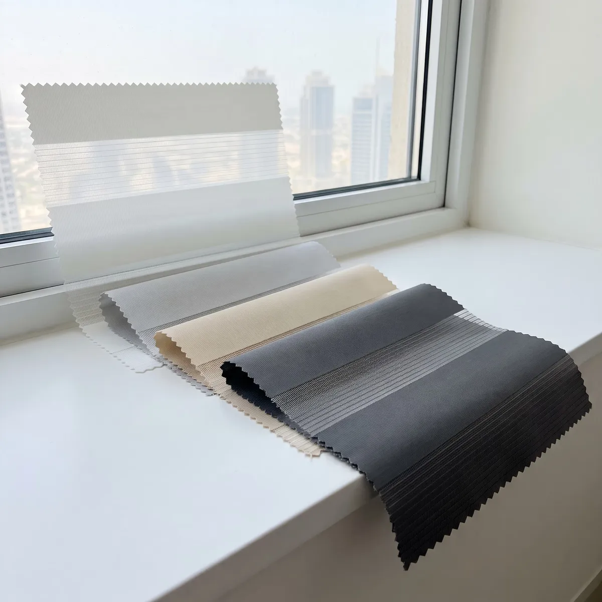 Zebra blind stripe samples with pinked edges on a sunlit sill, white, light grey, sand and charcoal, towers through the window