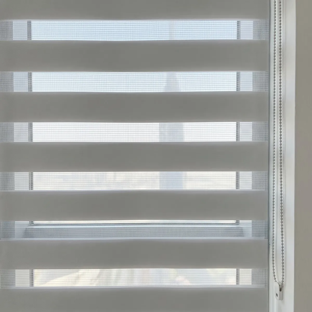 Light grey zebra blind fabric close up with its stripes half open, daylight showing through the sheer bands, a bead chain at the right